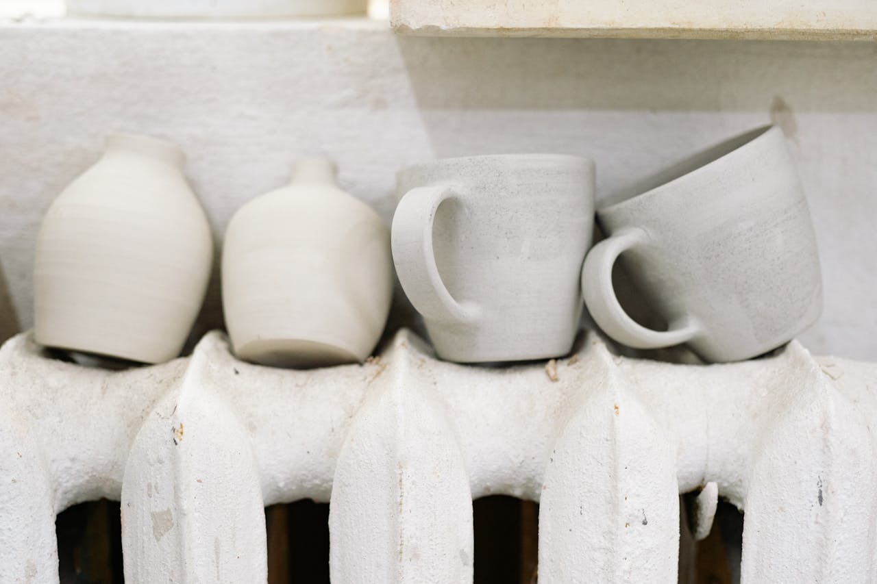 Handcrafted ceramic mugs and vases drying on a radiator in a pottery studio.