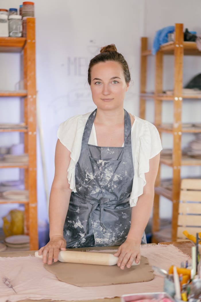 A woman in an apron crafting pottery with a rolling pin in a workshop.